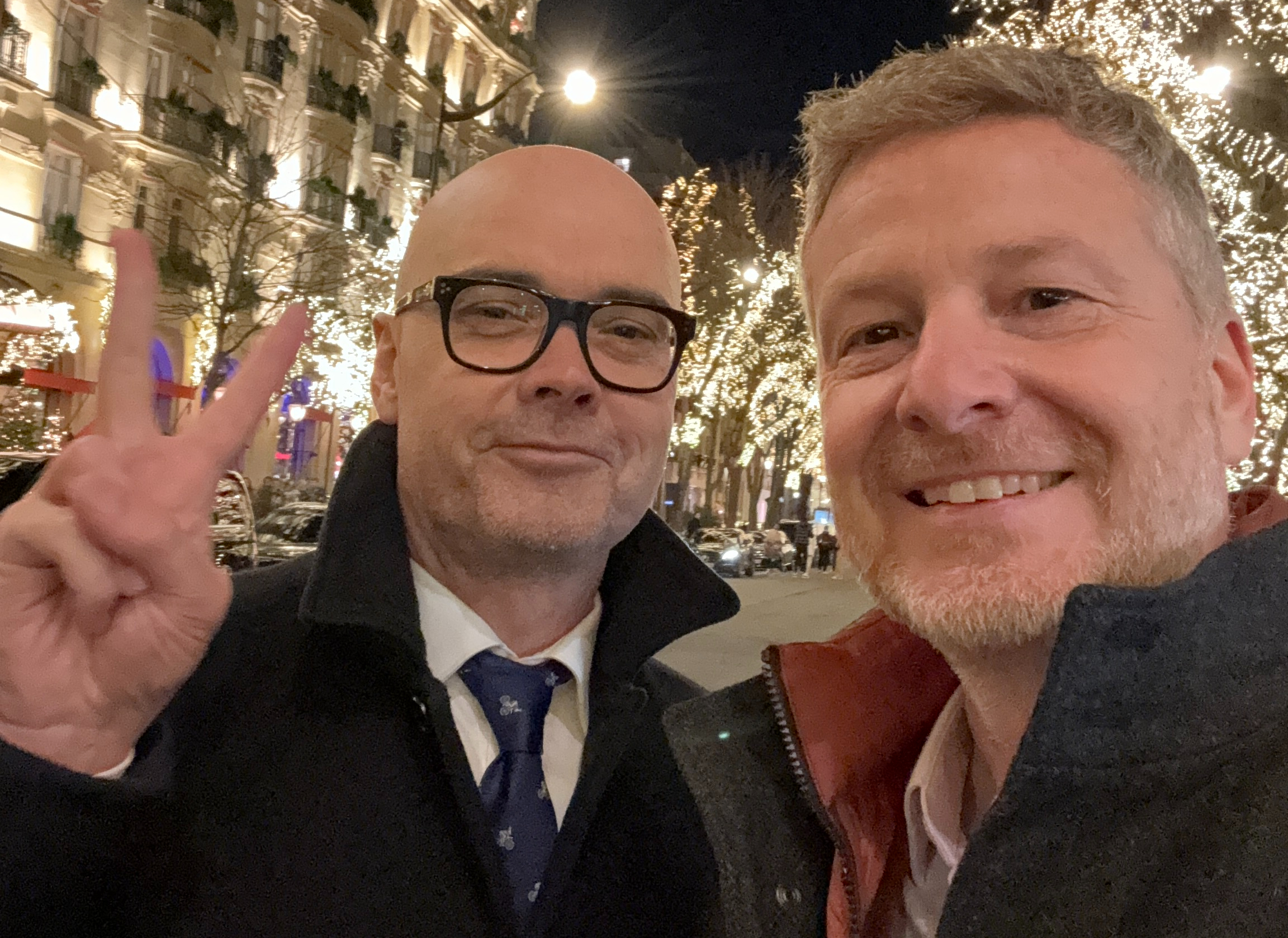 Two men smiling at the camera in a city scape filled with bright fairy lights